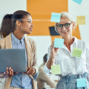 Sharing ideas. Two female coworkers putting colorful sticky notes on a glass wall and smiling while standing in the modern office, business people working together